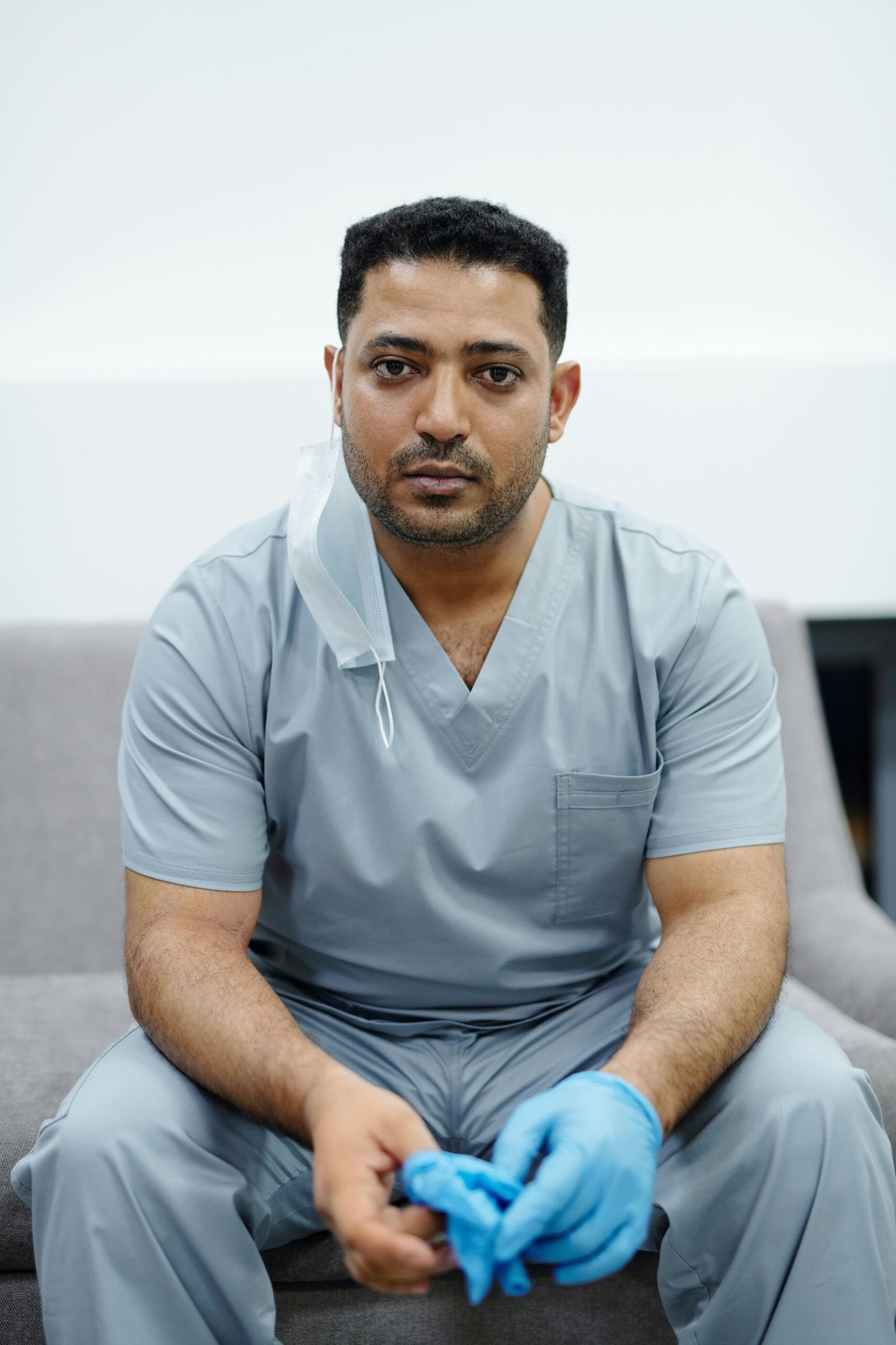 Healthcare professional seated on a couch in scrubs, wearing blue gloves, looking at camera.